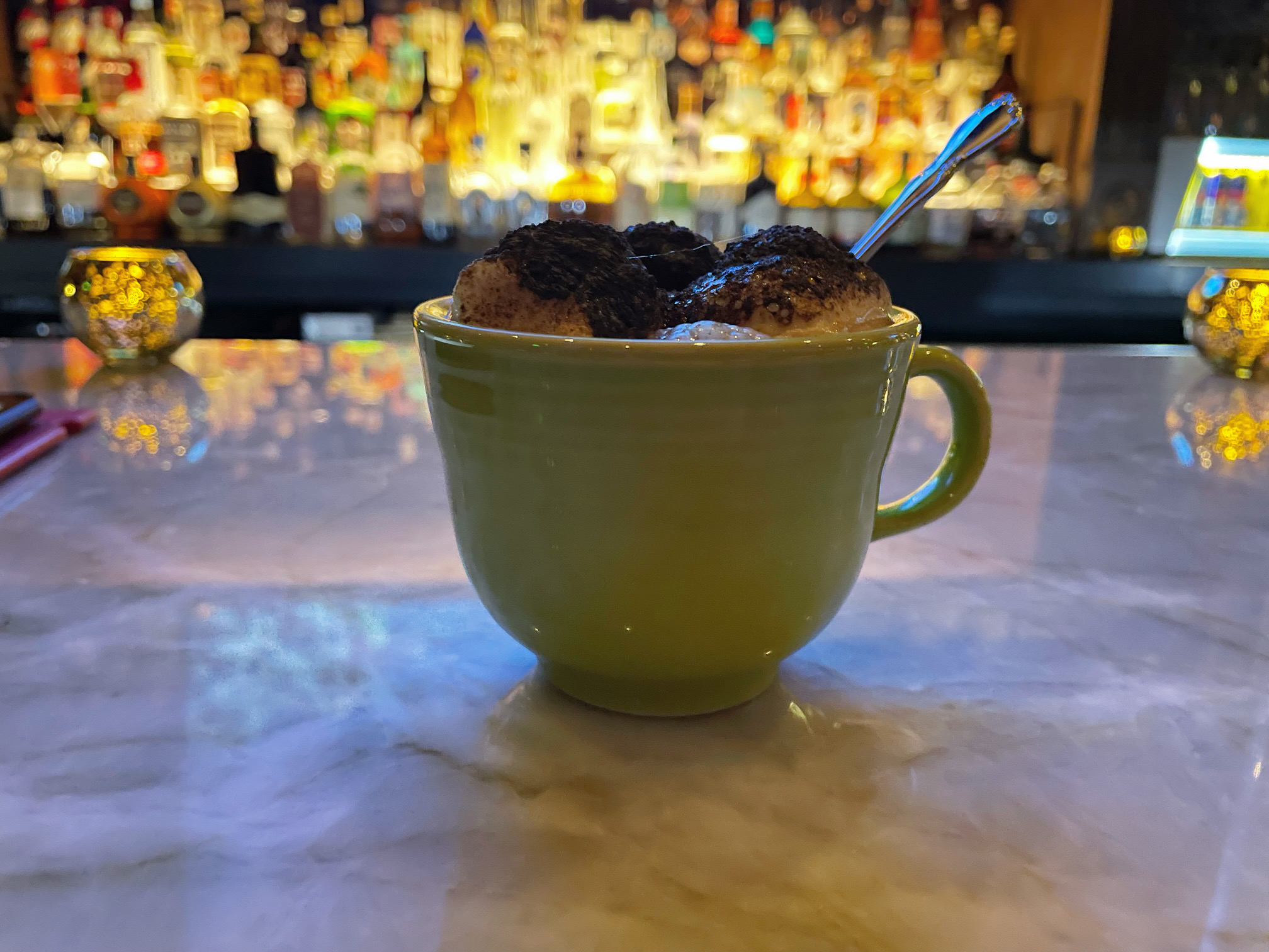 In a dimly lit bar, there is a green mug with charred marshmallows on top. Behind the drink is a bar with illuminated liquor bottles. Photo by Alyssa Buckley.