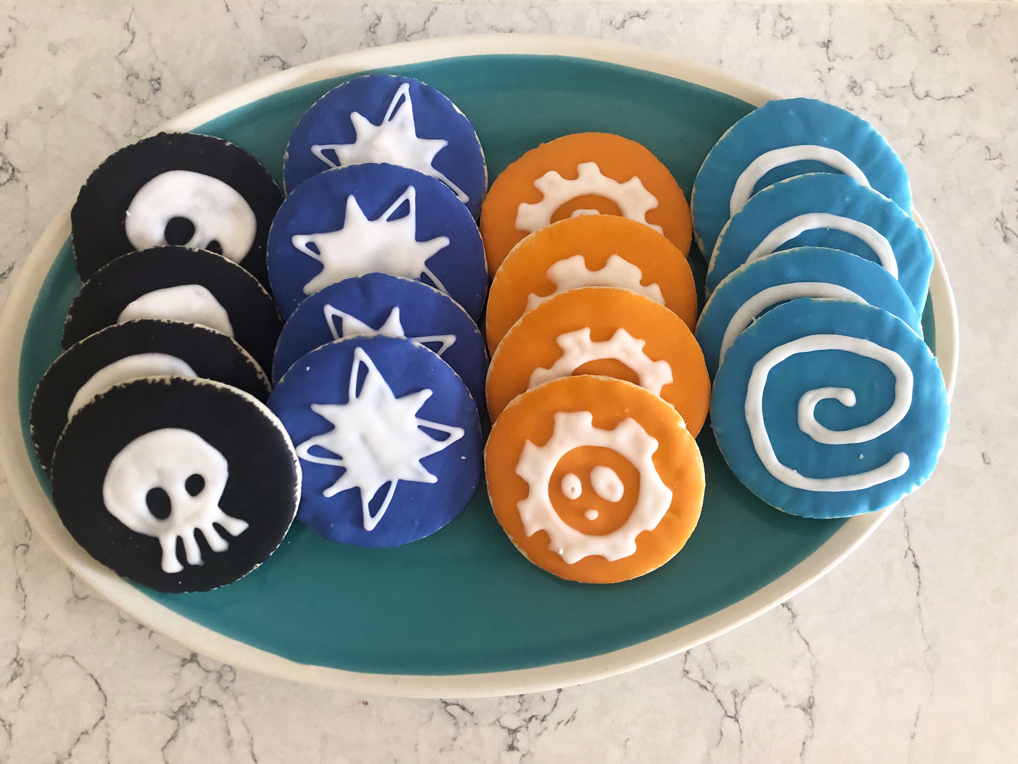On a teal plate, there are sixteen cookies. Four black with a skull, four purple with a star, four orange with a gear, and four blue with a swirl. Photo by Alyssa Buckley.