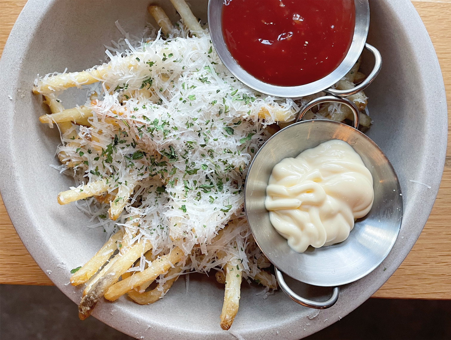 An overhead photo shows a gray bowl filled with skinny fries covered in Parmesan grated cheese with two little cups of sauce: one red and one white. Photo by Alyssa Buckley.