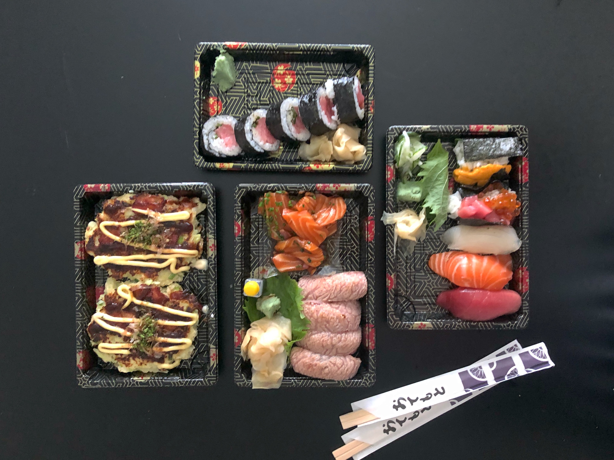An overhead photo of takeout rectangular dishes on a black table with two chopsticks packs. There are okonomiyaki, five piece nigiri, seared toro, marinated salmon, and a tuna sushi roll. Photo by Alyssa Buckley.
