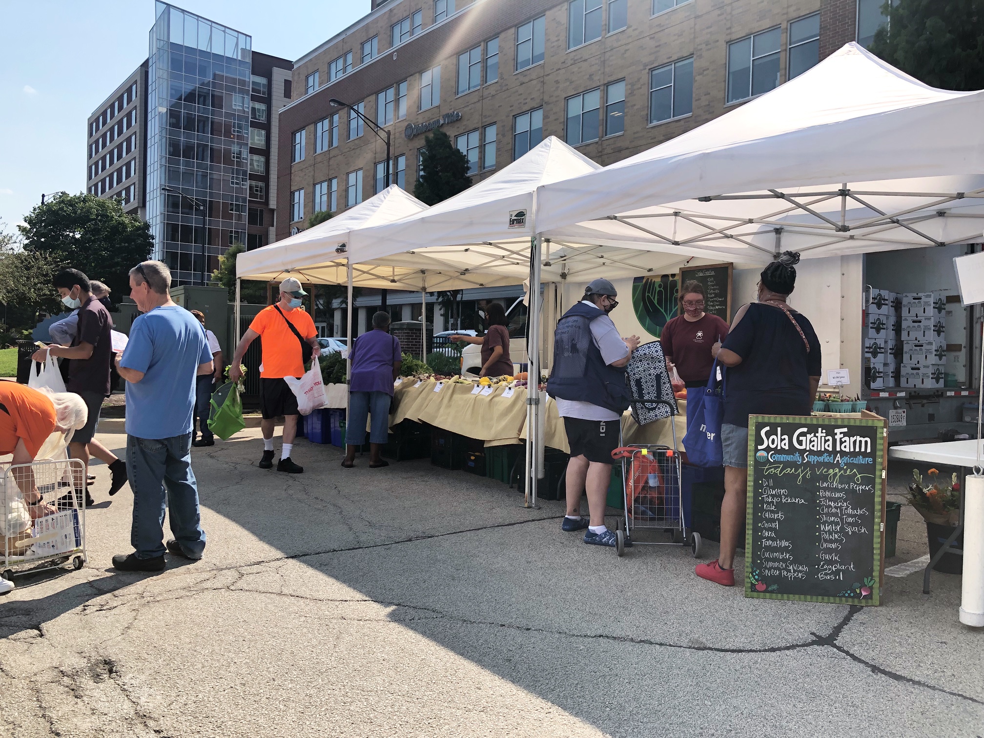 In the parking lot, the Champaign Farmers' Market vendors have tables under tents offering goods for sale. Masked and unmasked shoppers walk by, browse, and pay for goods with the Champaign skyline behind the tents on the right side of the photo. Photo by Alyssa Buckley.