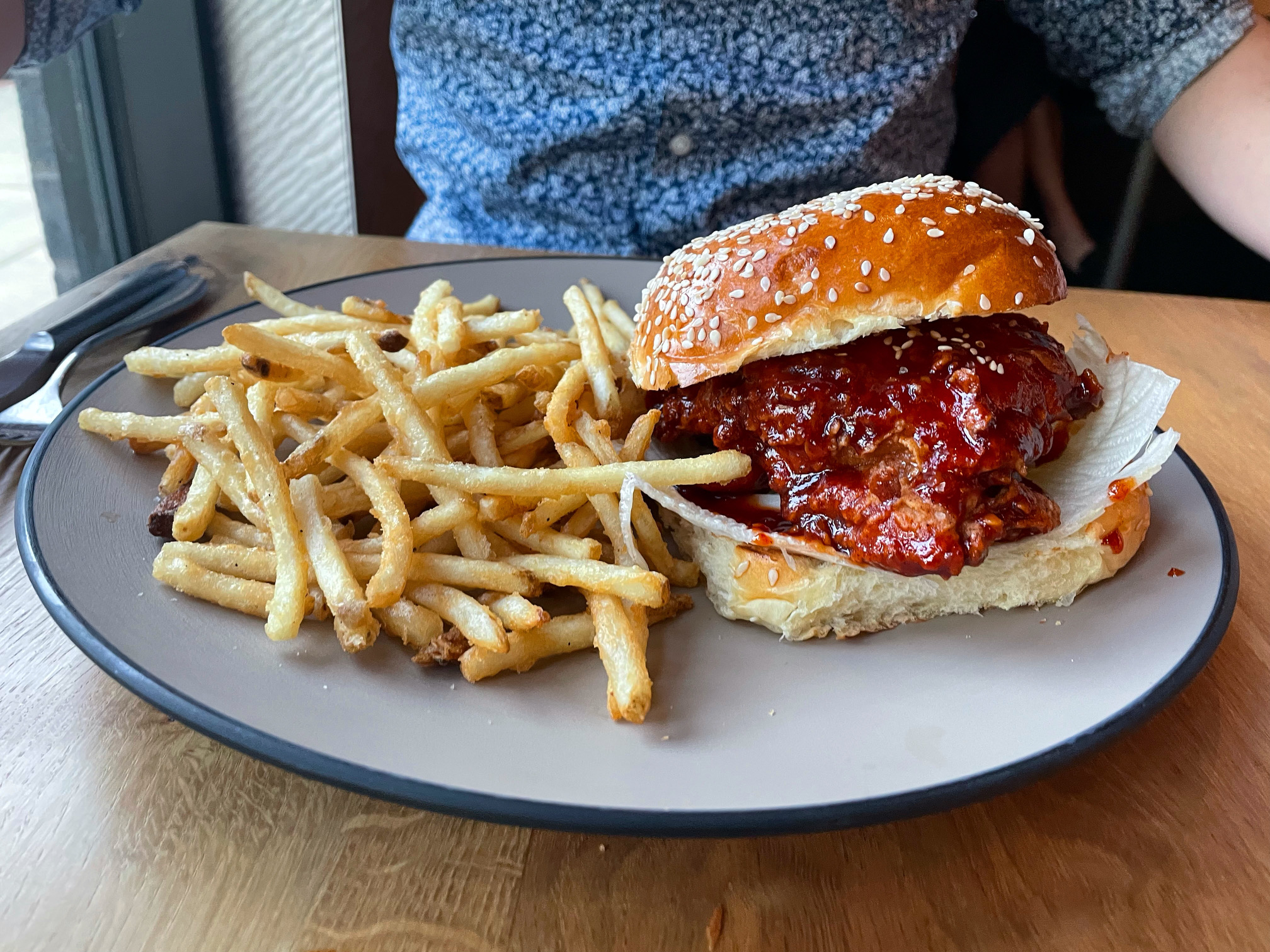 On a light wooden table, there is a gray plate with a big side of skinny fries beside a saucy Korean fried chicken sandwich on a sesame bun. Photo by Alyssa Buckley.