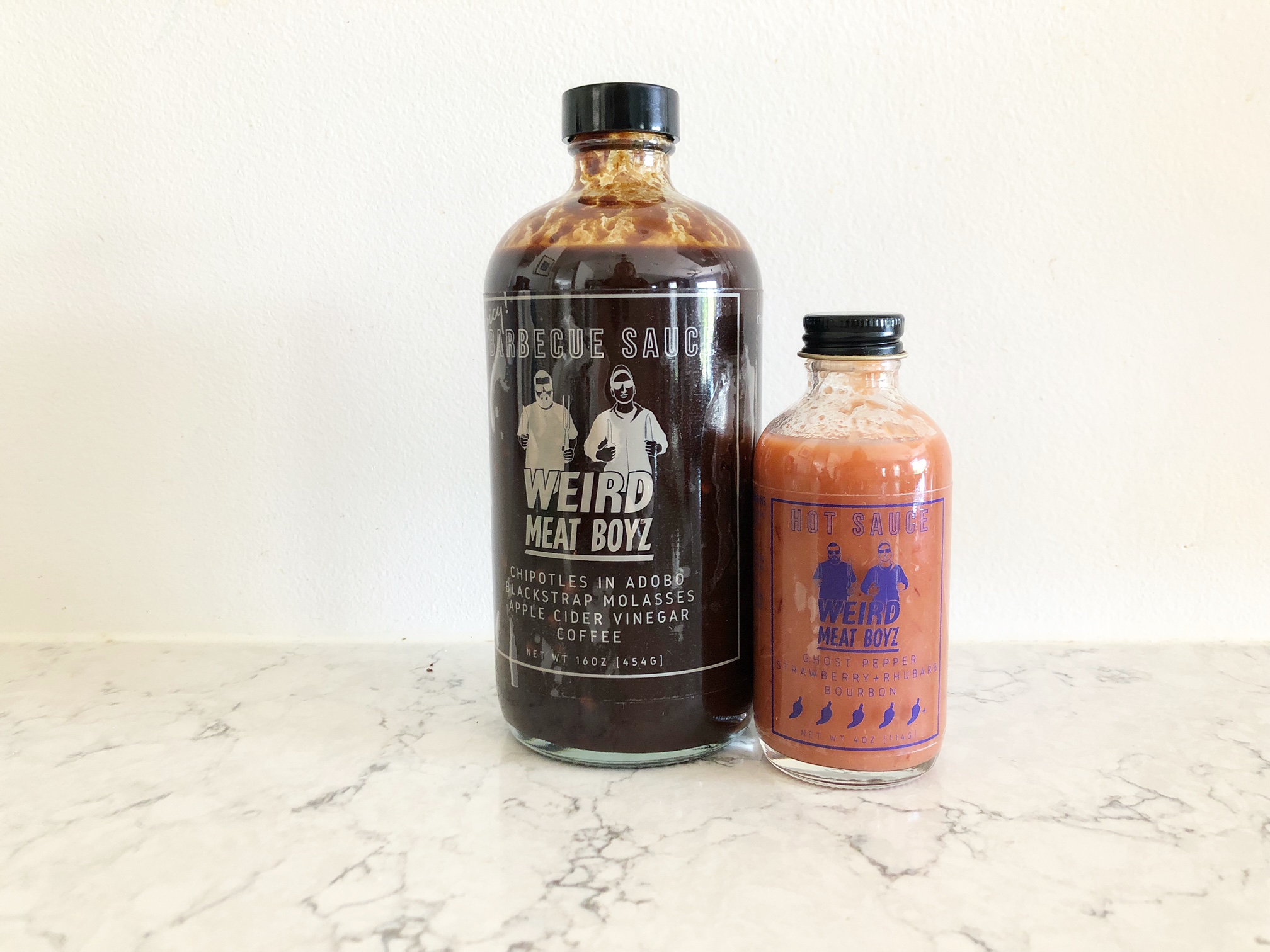 On a white counter in front of a white wall, there are two glass bottles of sauce. One is the barbecue sauce and the other is the ghost pepper sauce by Weird Meat Boyz. Photo by Alyssa Buckley.