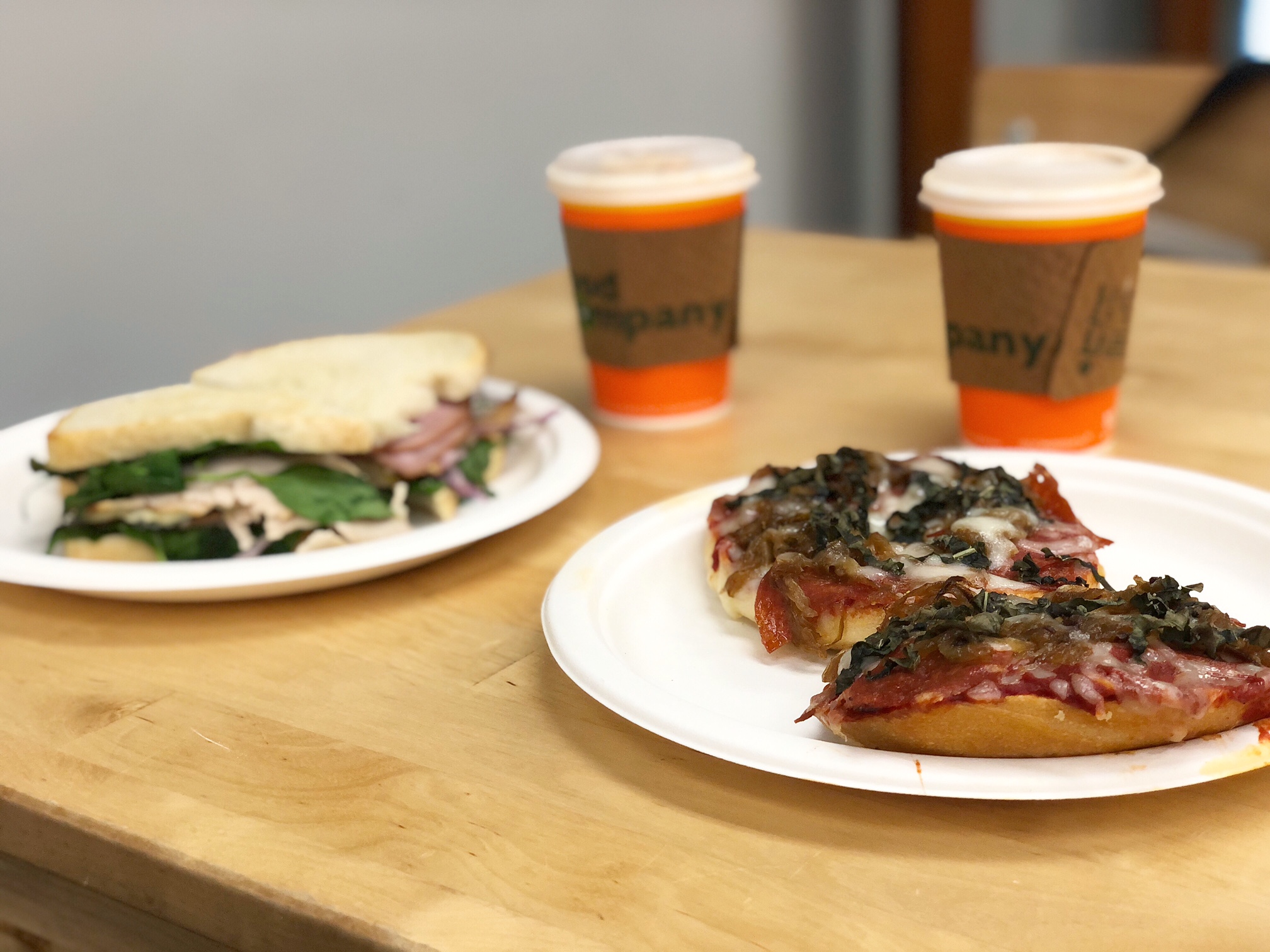 On a light wooden table, there is a sandwich on white bread cut diagonally in half and a pizza on another white plate. Behind the two plates are two hot coffee drinks in to go orange paper cups. Photo by Alyssa Buckley.