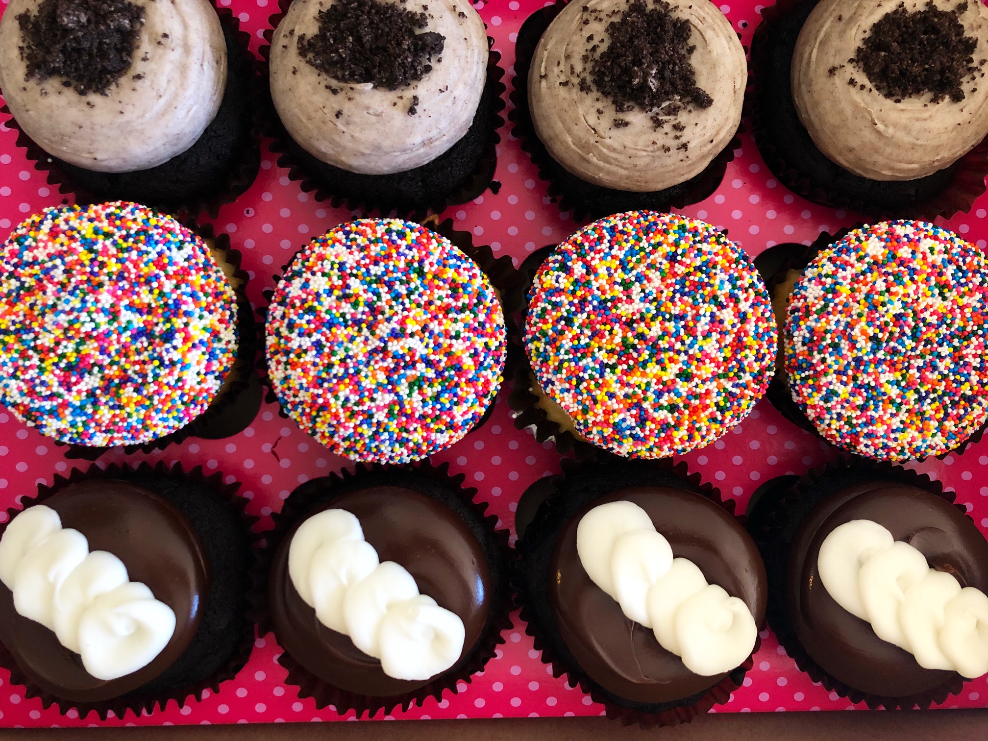 In a pink box, twelve cupcakes are lined up in three rows of four. The top row has gray frosting with black cookie crumbles. The middle row has white frosting with rainbow sprinkles, and the bottom row has black fudge frosting with a white diagonal line of frosting on top. Photo by Alyssa Buckley.