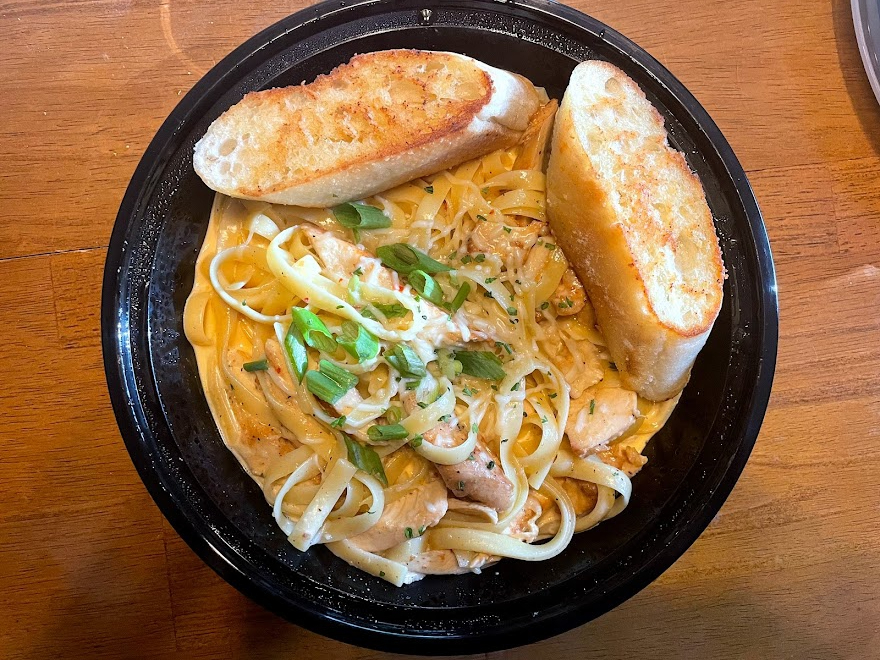 On overhead photo shows the chicken alfredo dish from PastaMania food truck with a slice of garlic bread on top of alfredo pasta in a black circular takeout container. Photo by Remington Rock