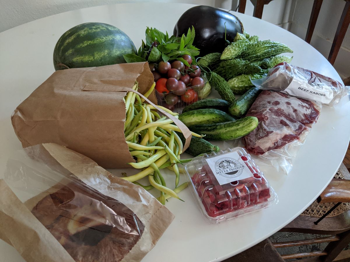 n the forefront of the picture, we can see a plastic and brown paper package with a danish inside and a clear plastic carton with red raspberries. Behind these two items, are light green beans spilling out of a brown paper bag, pickling cucumbers, beef kabob meat in vacuum-seal packages, bittermelon, cherry tomatoes, green leafy Thai basil, a dark shiny eggplant, and a striped green watermelon. All the food sits atop a round white table. Photo by Tias Paul.