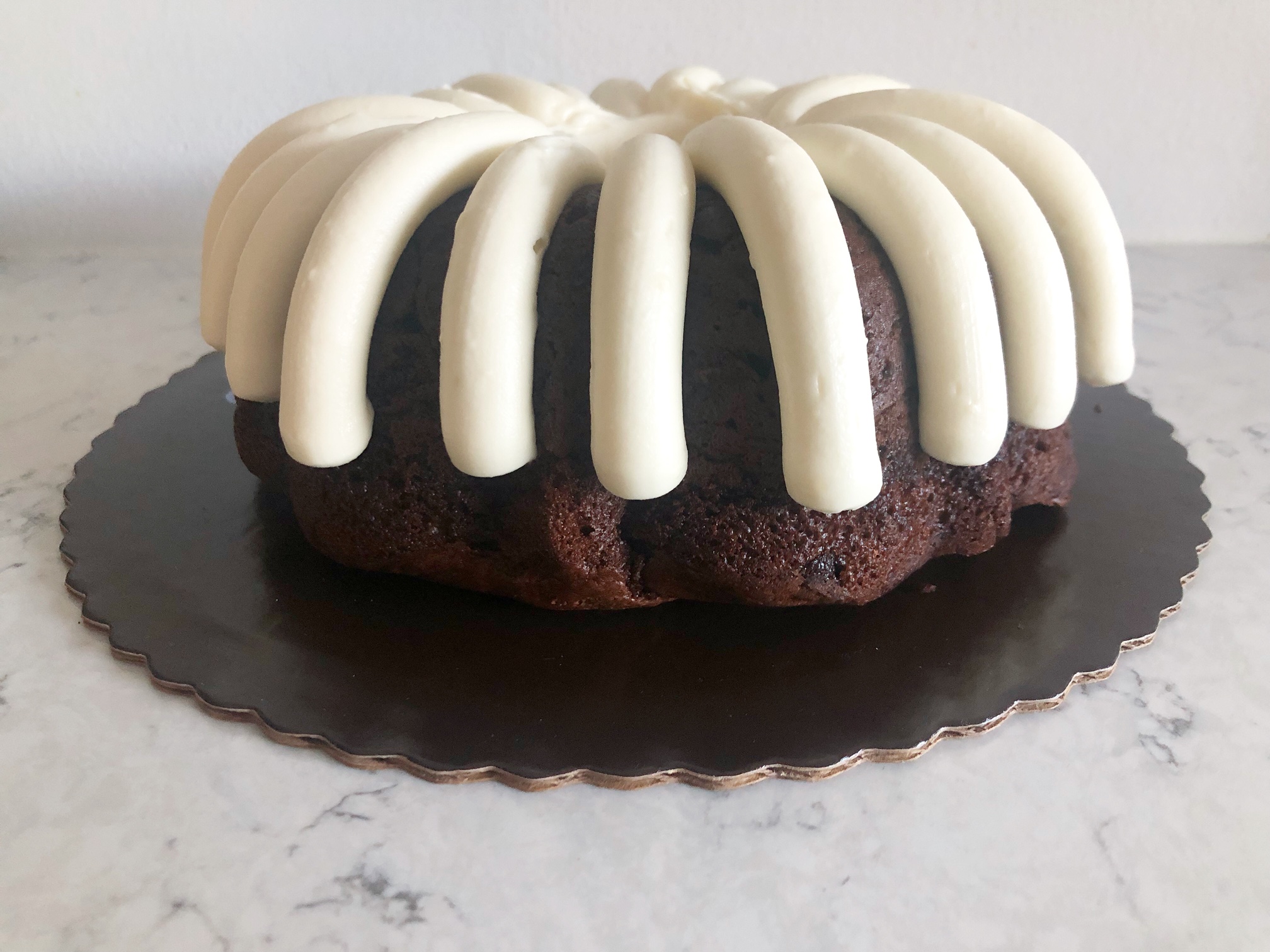 On a marble counter, there is a scalloped cake plate with a chocolate cake and signature cream cheese frosting stripes from Nothing Bundt Cakes. Photo by Alyssa Buckley.