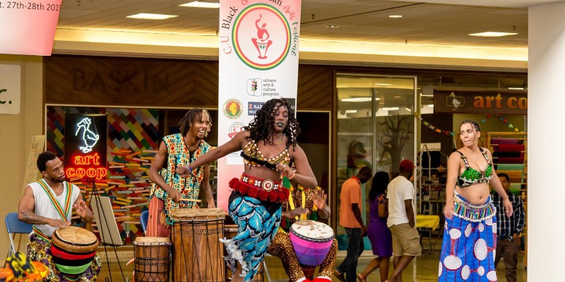 Two women in brightly colored African regalia are dancing in front of a row of drummers. Photo from C-U Black and African Arts Festival. 