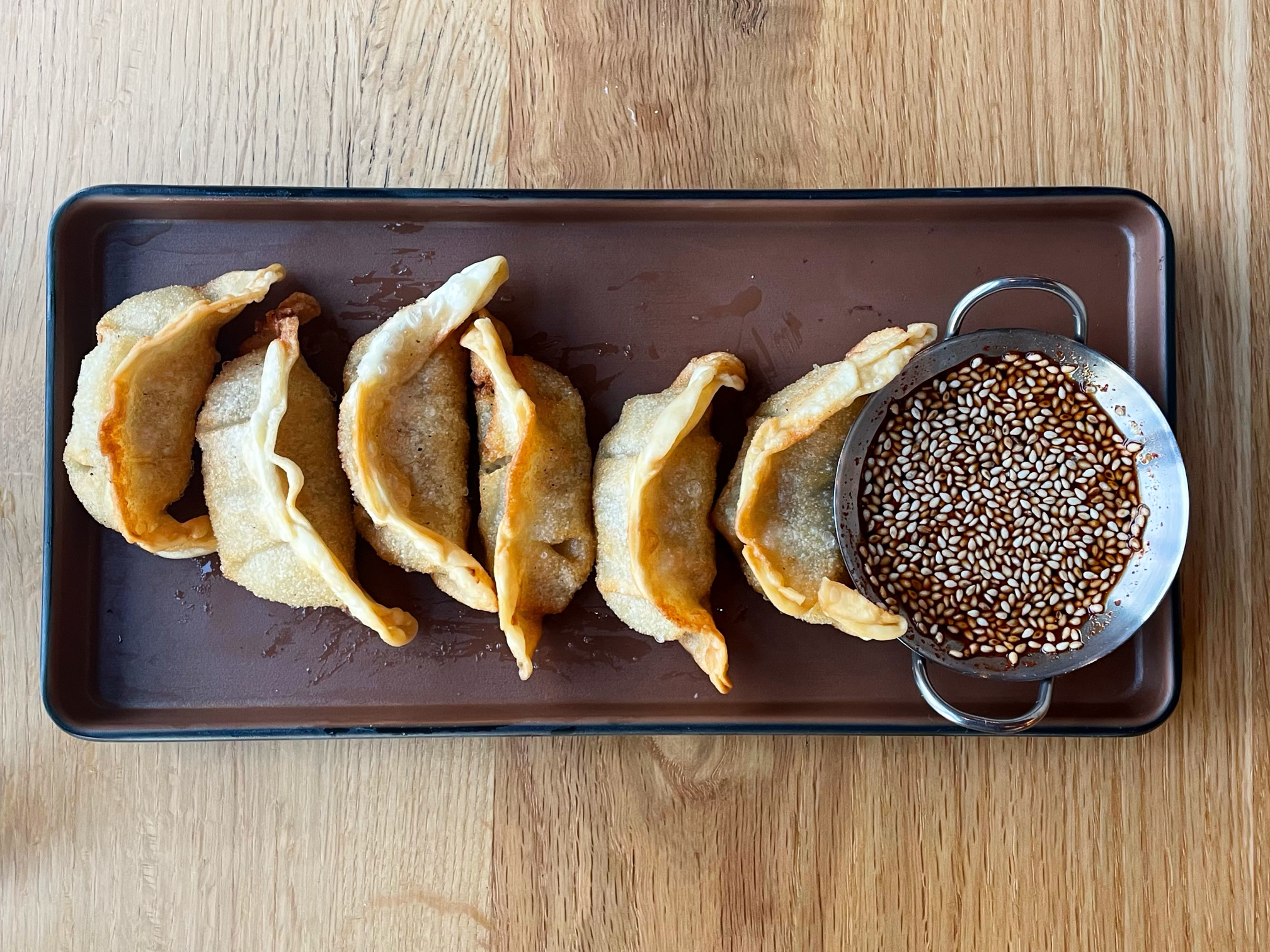 An overhead photo shows a rectangular brown plate with six fried dumplings. On the right, there is a small, miniature wok filled with a brown sauce and lots of sesame seeds. Photo by Alyssa Buckley.