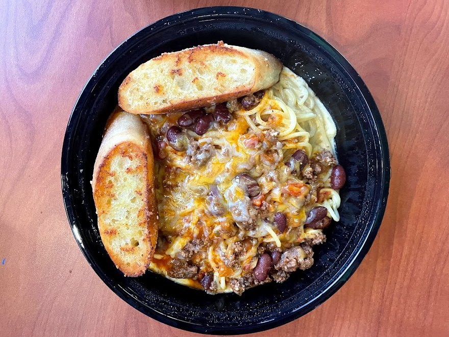 On overhead shot, there is a black circular takeout container with chili mac pasta and a slice of garlic bread. Photo by Remington Rock.