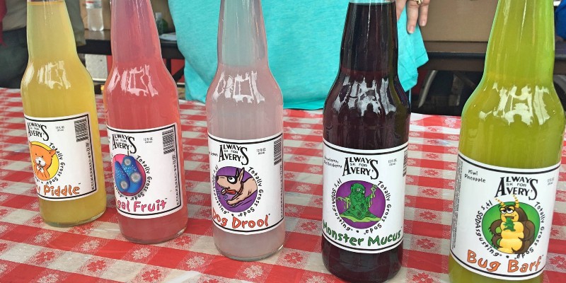 A row of brightly colored sodas in glass bottles. They are lined up on a red and white checked table cloth. Photo by Ashley Huddson. 