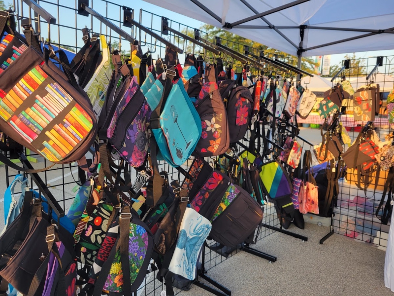 A view of Kobakâ€™s bag display at Urbanaâ€™s Market at the Square. Colorful bags hang on black racks and hooks against a white tent background. Photo by Kate Kobak.