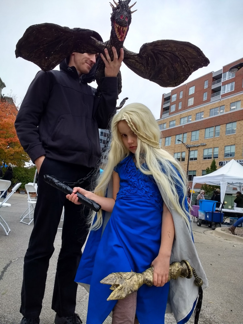 Kobakâ€™s daughter and husband in Halloween costumes. Her daughter is dressed as Daenerys Targaryen in a royal blue dress and cape, long white wig with braids, and leggings with brown boots. Kobakâ€™s husband is dressed in all black holding a paper mÃ¢chÃ© dragon attached to a chain, which the daughter is holding. Photo by Kate Kobak.