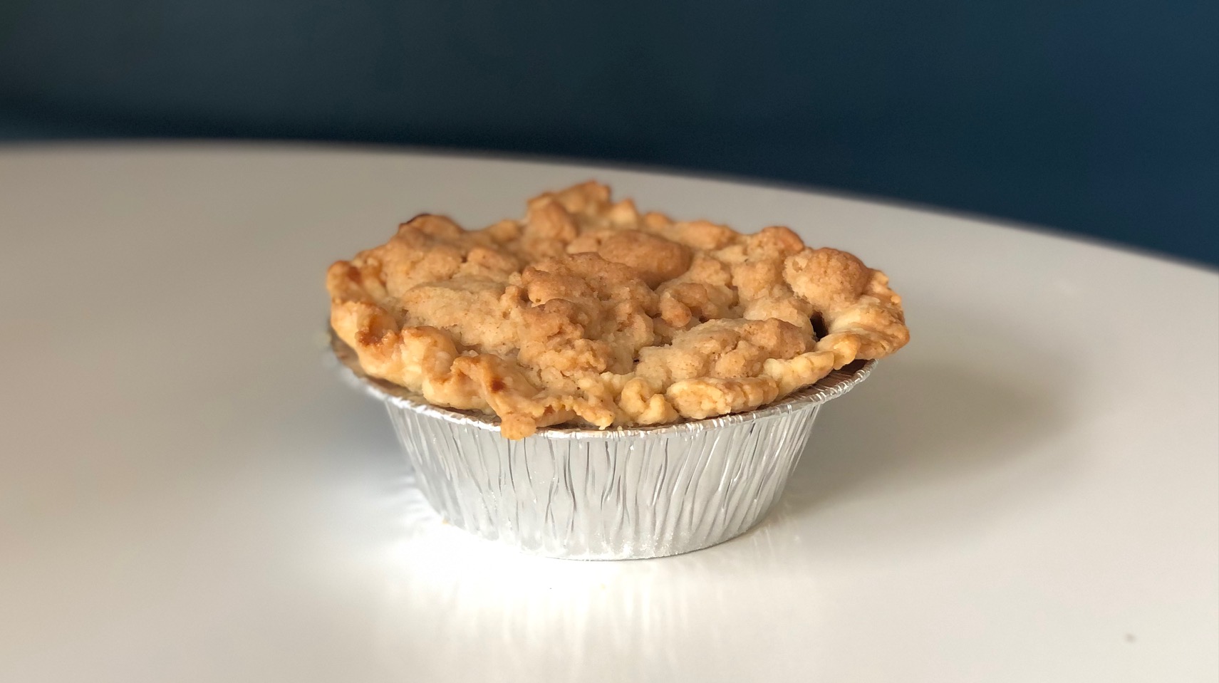 On a white table, there is a mini apple pie with a crumble topping in a tin foil pan. Photo by Alyssa Buckley.