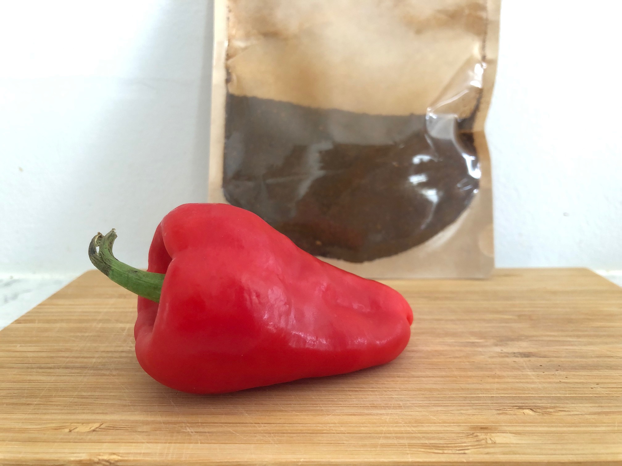 On a wooden cutting board, a red paprika pepper lays uncut on its side. Behind it is a plastic bag of dried paprika spice. Photo by Alyssa Buckley.