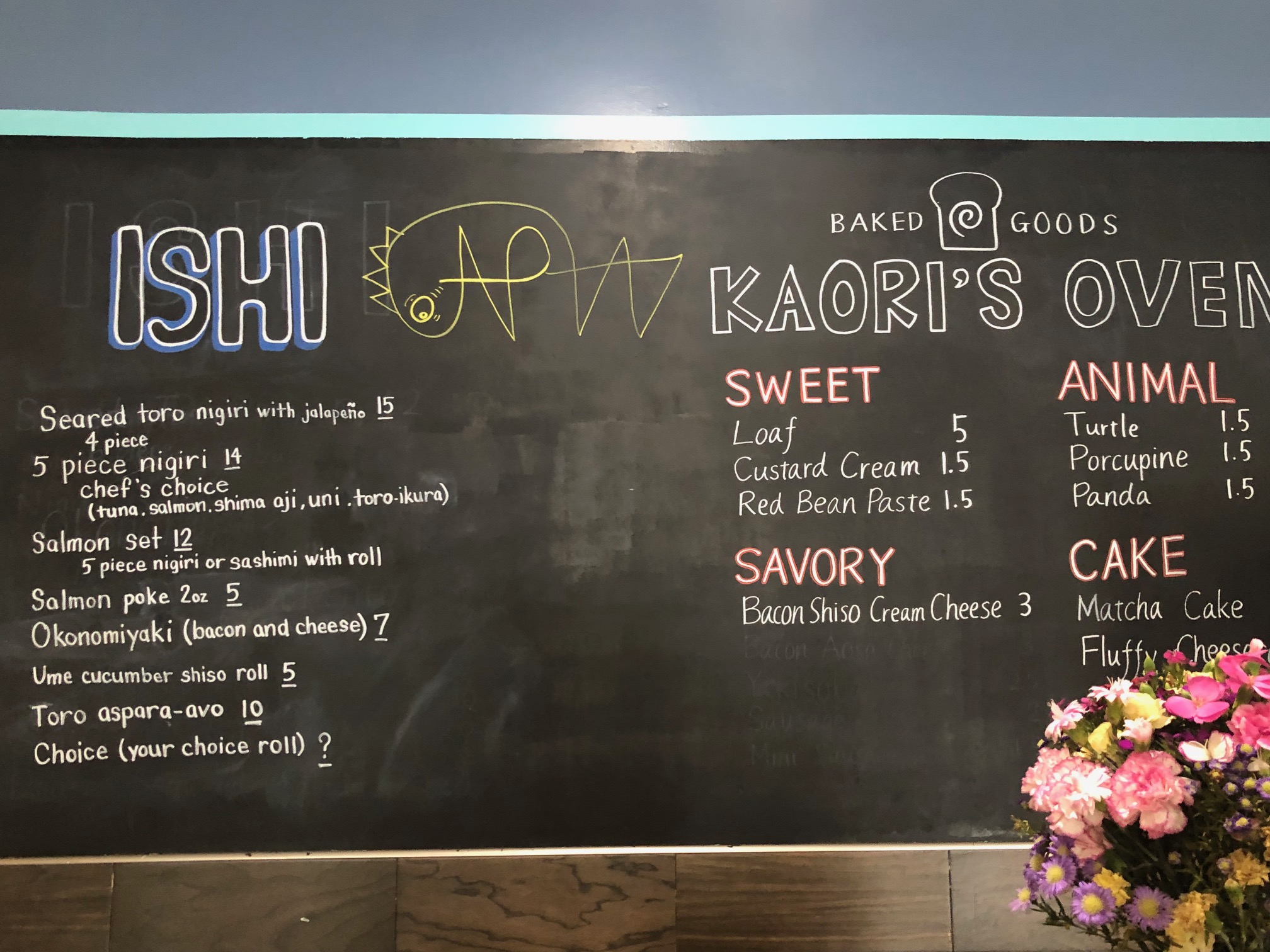 On a large black chalkboard, there are two sides of available options. On the left, there is ISHI and a handwritten list of sushi dishes and the price. On the right side is Kaori's Oven which has desserts listed by Sweet, Savory, or Animal. Photo by Alyssa Buckley.