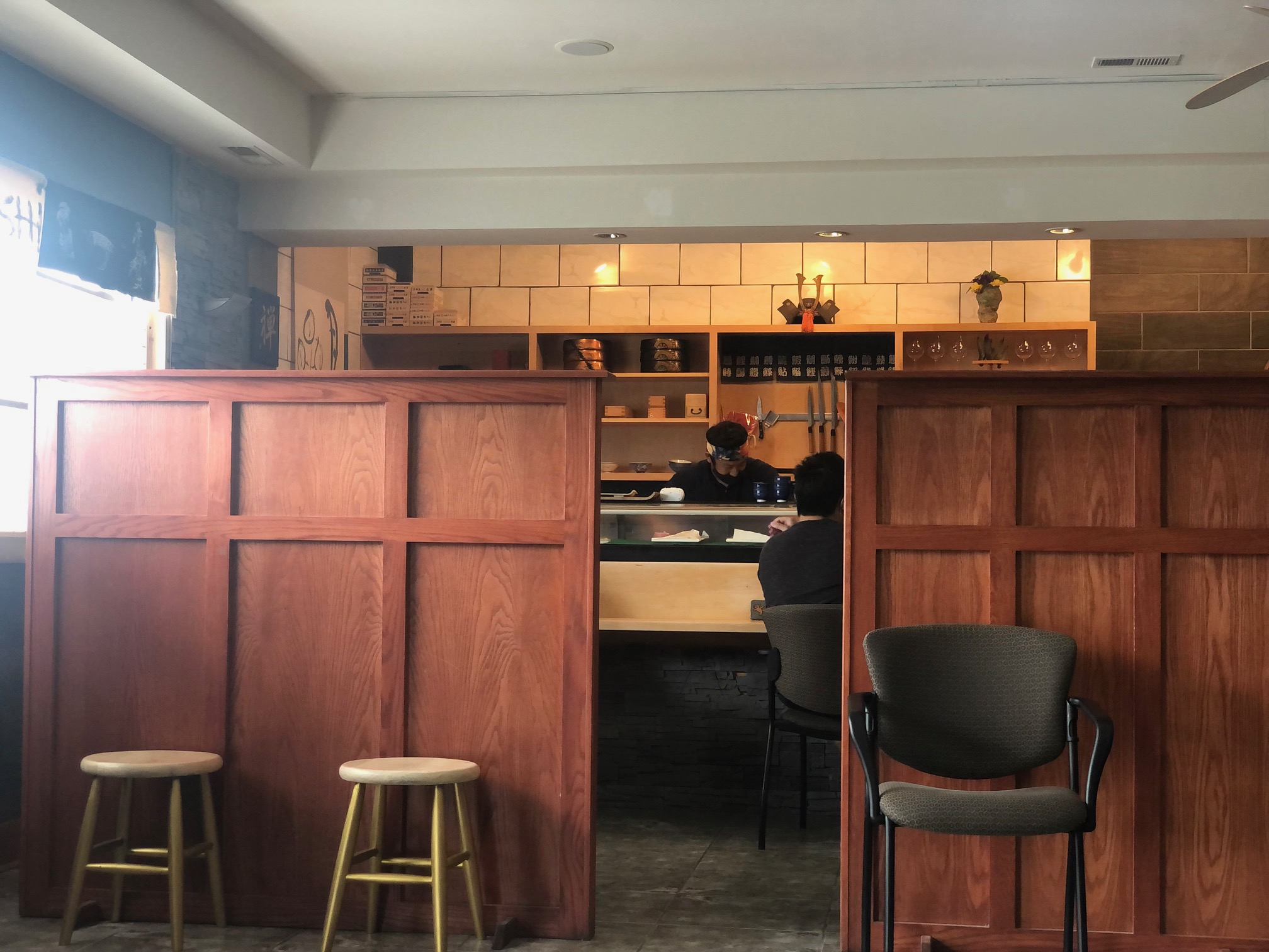 Inside ISHI, there is a sushi bar beyond wooden dividers. The front room has two wooden stools and a black office chair with no one sitting in them. Photo by Alyssa Buckley.