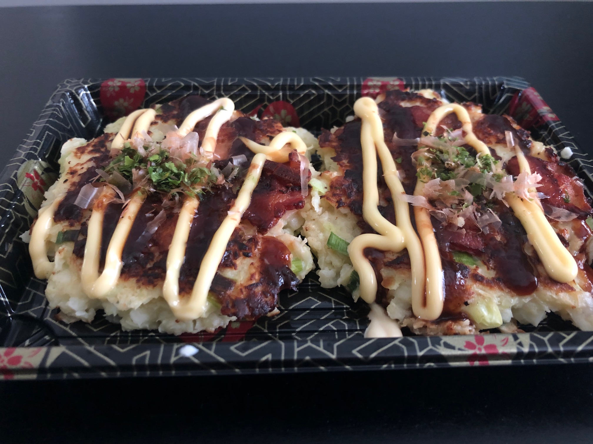 Two okonomiyaki are in a rectangular container with drizzles of yellow and dark brown sauces. Photo by Alyssa Buckley.