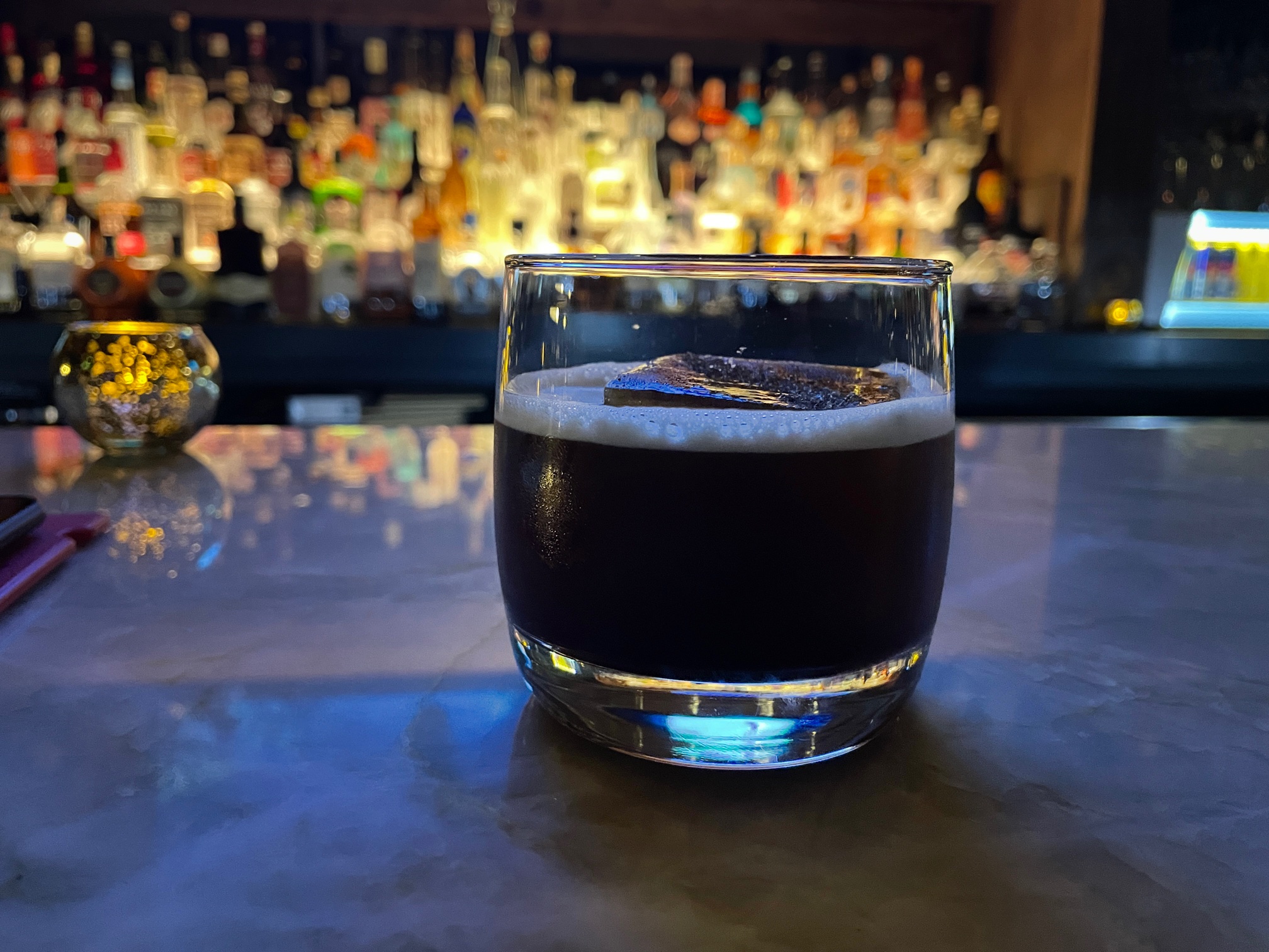 In a dim bar, there is a half glass of a dark cocktail with a large ice cube in it. Behind the glass is the bar's bottles of liquor lit up from underneath. Photo by Alyssa Buckley.