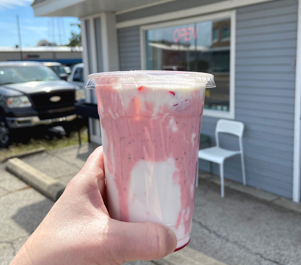 A to-go beverage held up by a white personâ€™s hand in front of Tasty Tart in Champaign, IL. The beverage is white with pink drizzles inside of the plastic cup. Photo by Jessica Hammie. 
