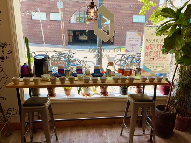 Inside the campus location of Brew Lab, there is a long, skinny table in front of a window with many white coffee mugs on it, ready for the author's coffee cupping experience. Photo by Stephanie Wheatley.