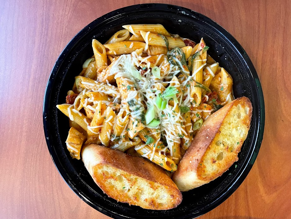 An overhead photo of the inferno pasta shows a black circular takeout container on a wooden table. In the takeout container, there is penne pasta with shredded Parmesan on top. Photo by Remington Rock.
