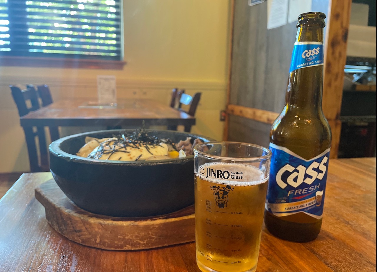 On a wooden table, there is a black stone bowl with bibimbap and Cass beer poured into a beer glass. Photo by Alyssa Buckley.