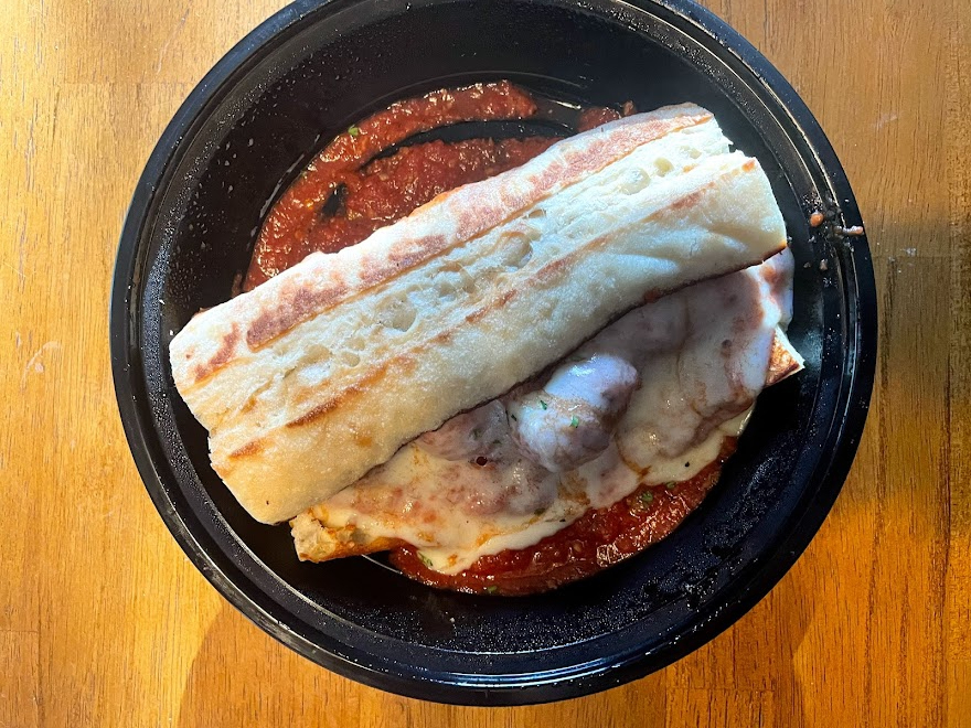 On a wooden table, there is a meatball sub sandwich in a black takeout container on a wooden table. Photo by Remington Rock.