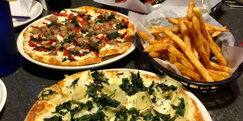 There are two pizzas, uneaten, and a basket of fries on a dark table. Photo by Sarah Meilike.