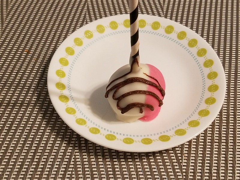 A strawberry cake pop standing on end on a small plate with a stick length more than five times the diameter of the cake pop. Photo by Matthew Macomber.