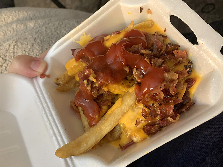 In a small square styrofoam container held by a white hand in front of a light blue fleece blanket, there are fries topped with orange cheese and bacon crumbles. Photo by Zoe Valentine. 