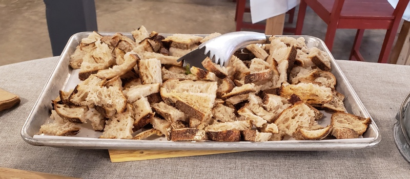 An image showing a large silver cookie sheet lined with parchment paper and filled with sliced bread. There is a silver pair of tongs resting on the food. Photo by Sara Ressing.