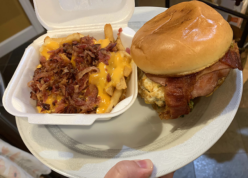 On a white plate held by a white person's hand, there is a styrofoam square container with cheese fries and beside it on the plate is a cheeseburger with bacon slices showing. Photo by Zoe Valentine. 