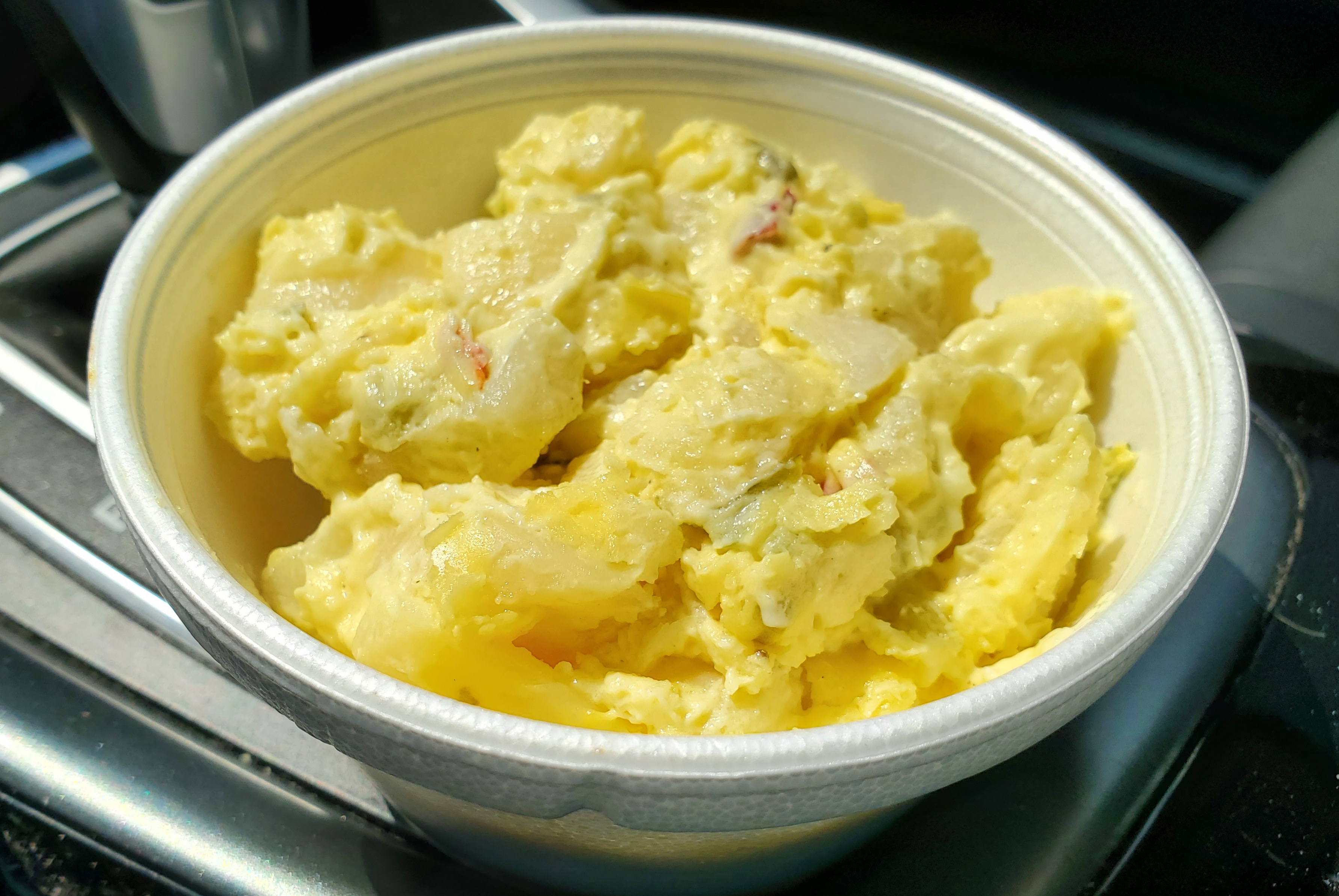 In a silver car, there is a white styrofoam bowl full of yellow potato salad. Photo by Carl Busch.