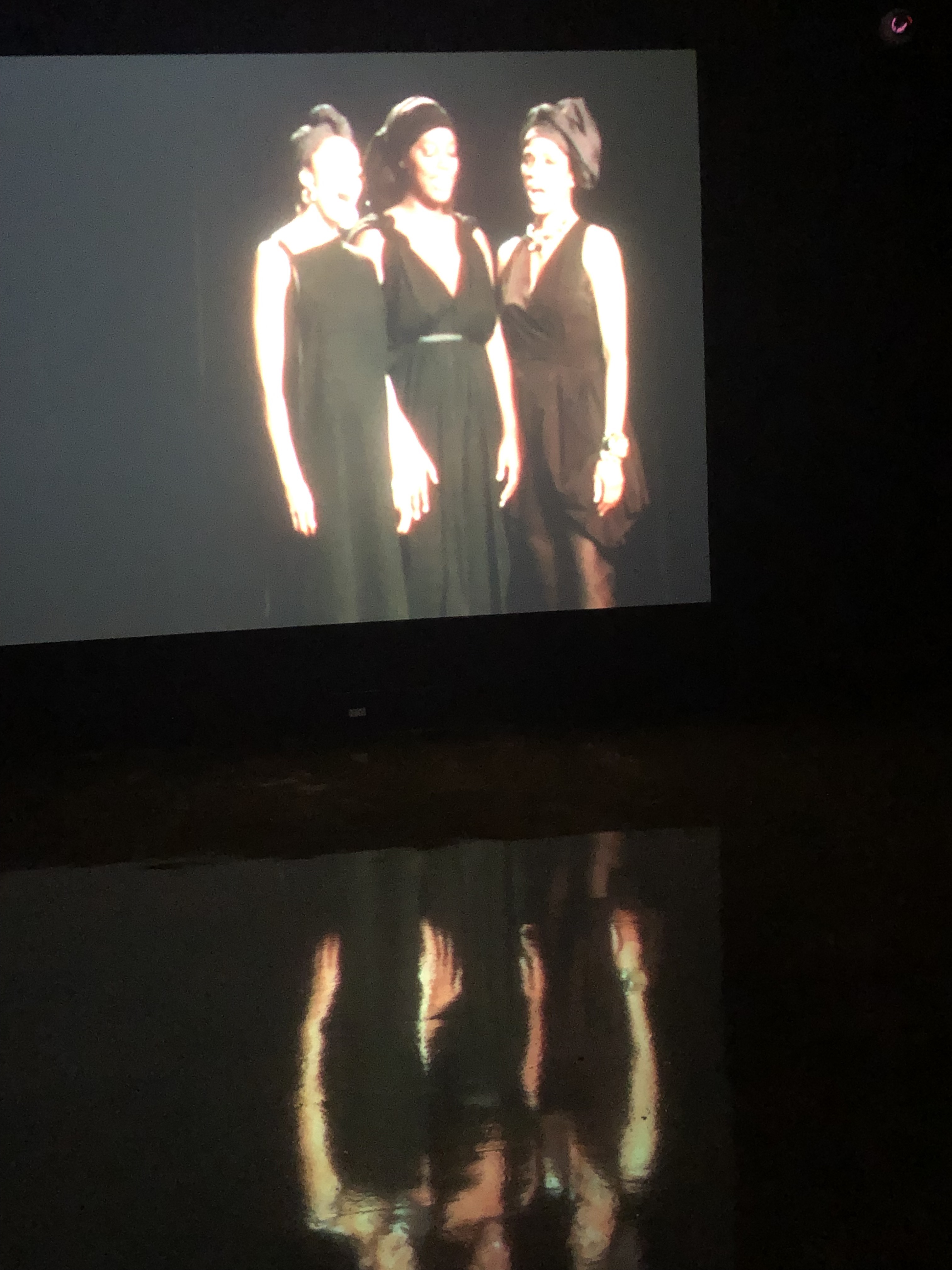 A scene from Carrie Mae Weemsâ€™ film in which three Black women are singing in harmony in elegant black dresses against a stark black backdrop. Photo by Amy Penne.