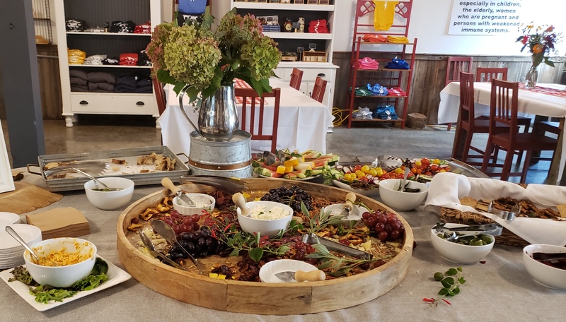 This is a view of the dining area at Prairie Fruits Farm & Creamery. There are tables with red chairs, white table cloths and red checkered runners. There are shelves against the back wall with logo clothing items available for purchase. In the foreground of this photo is an array of food items and appetizers offered for the event. They include bowls of olives, and cheese spreads, trays of bread, crackers, fruits and vegetables and a charcuterie with grapes, cheeses, dried fruit and nuts. Photo by Sara Ressing.