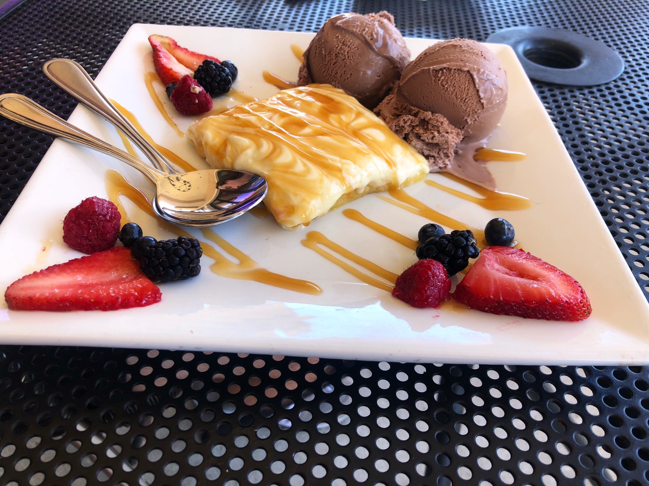 On a black metal outdoor table, there is a white square plate with a yellow gooey butter cake garnished with fresh berries and two scoops of chocolate ice cream. Photo by Alyssa Buckley.