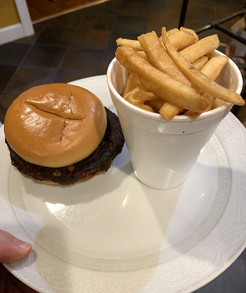 On a white plate held by a white person's hand, there is a burger beside a paper cup of fries. Photo by Zoe Valentine. 