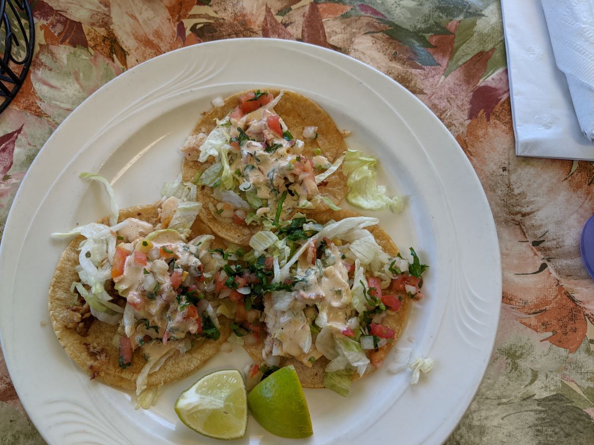 The tacos are made of yellow corn tortillas and a colorful mix of salsa and grilled tilapia. The tacos are on a white plate, which rests on a table with a floral tablecloth. Photo by Tias Paul.