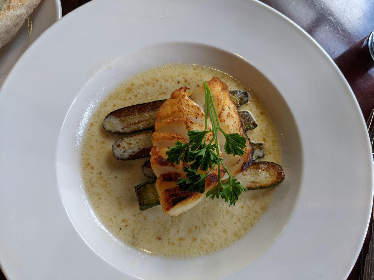 Golden pan-fried cod sits over green baby zucchini in a yellow cream sauce. The fish is plated in a wide-rimmed white bowl. The cod is garnished with a sprig of green parsley. Photo by Tias Paul.