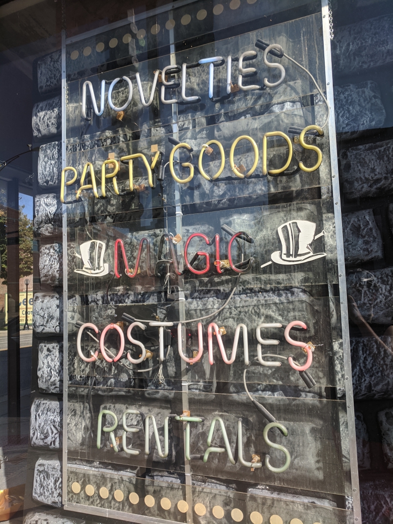 A close up of a window with neon signs reading horizontally: Novelties, Magic, Costumes, Rentals. Photo by Tom Ackerman.