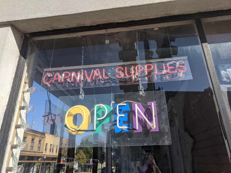 A window with two neon signs, one reading carnival supplies and one reading OPEN. Photo by Tom Ackerman.