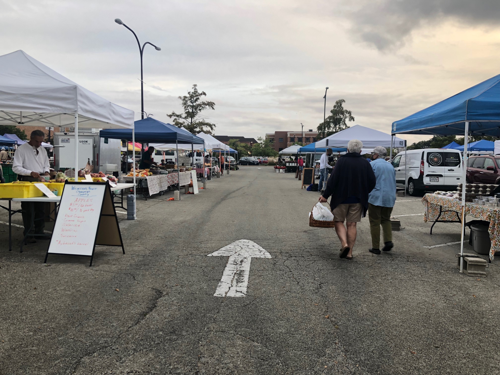 On a cloudy day, there are vendors under tents at the Urbana Market at the Square. The concrete has a weathered white arrow pointing away from the camera. Shoppers with bags face away. Photo by Alyssa Buckley.