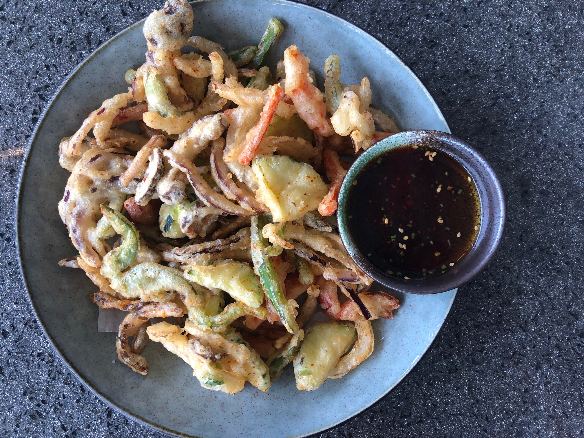 On a gray table, there is a white bowl of fried tempura vegetable strings with a side cup of soy sauce with sesame seeds. Photo by Alyssa Buckley.