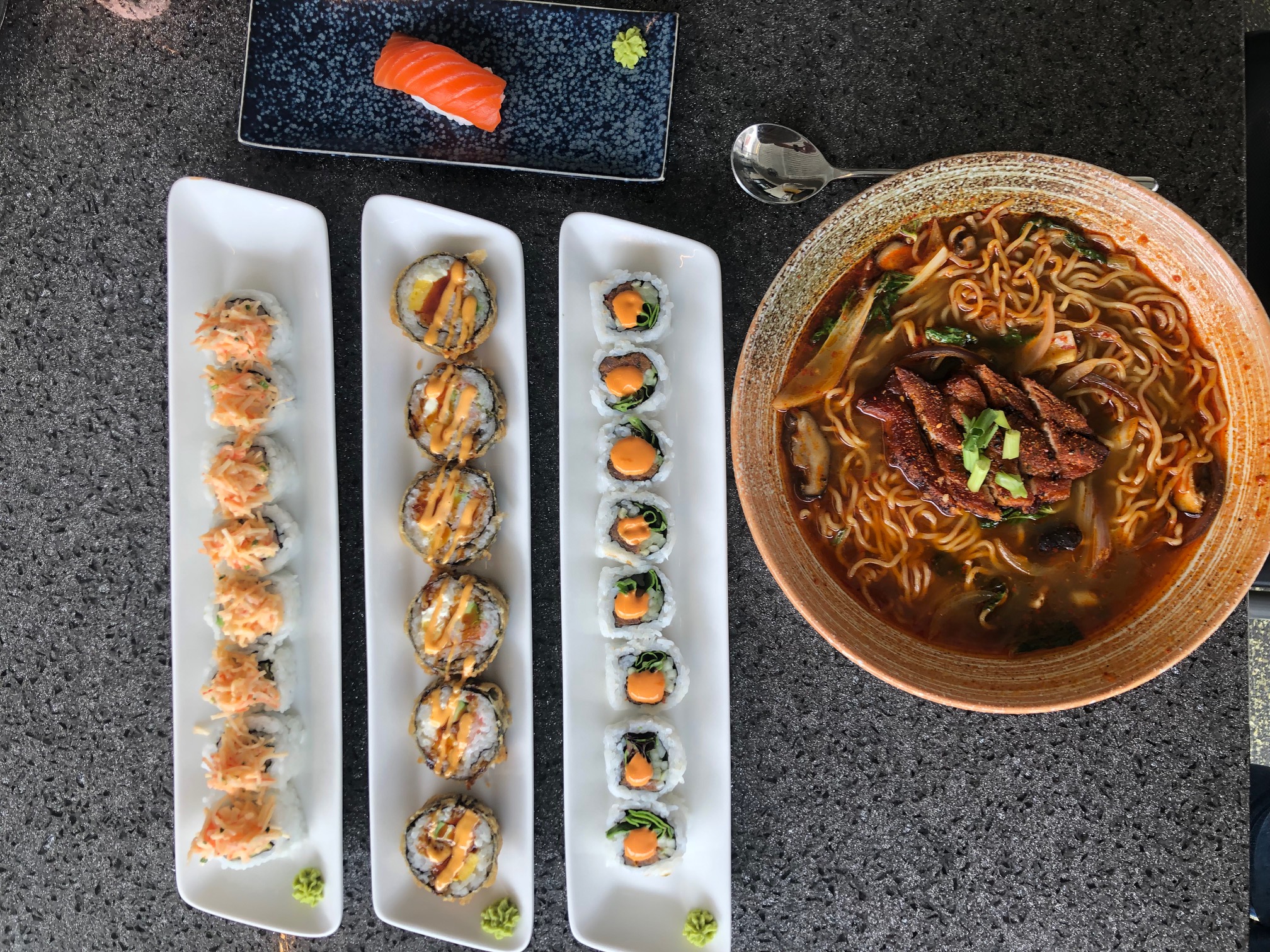 An overhead photo shows the author's lunch: pork belly ramen in a brown bowl, three sushi rolls each on their own white rectangular plate, and a small black plate holding nigiri salmon. Photo by Alyssa Buckley.