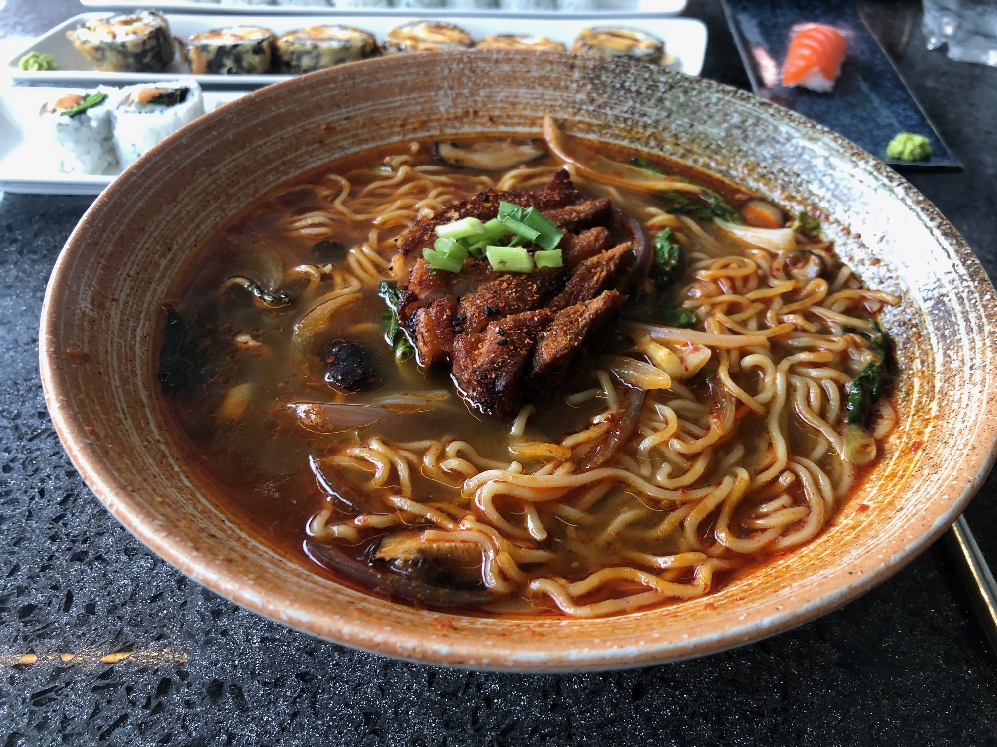 In a beige bowl, there is pork belly ramen. The ramen noodles float in a reddish broth with charred pork belly on top. Photo by Alyssa Buckley.
