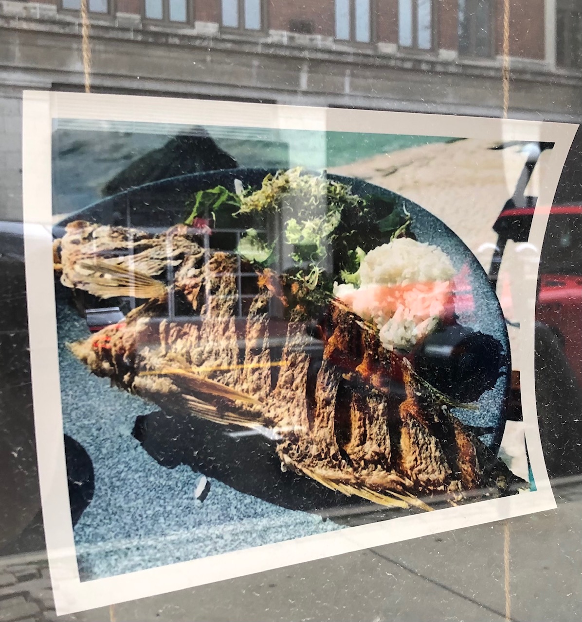 In a print out of a photo taped to the inside of a window, there is a cooked whole fish with slices on the gills on a black plate. Photo by Alyssa Buckley.