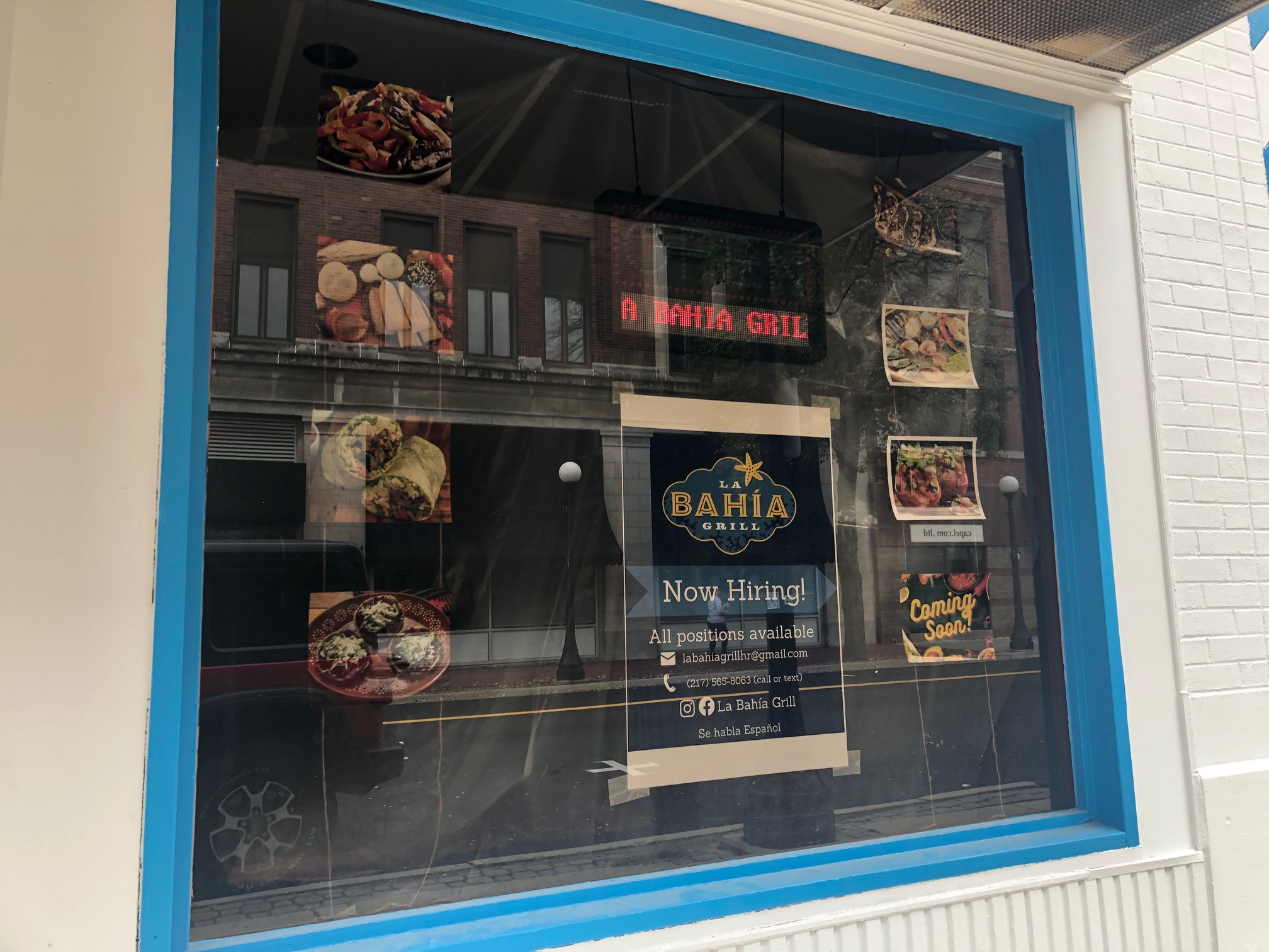 On brick painted white, there is a window with photos of food taped to the inside with a sign reading: La Bahia Grill Now Hiring. Photo by Alyssa Buckley.