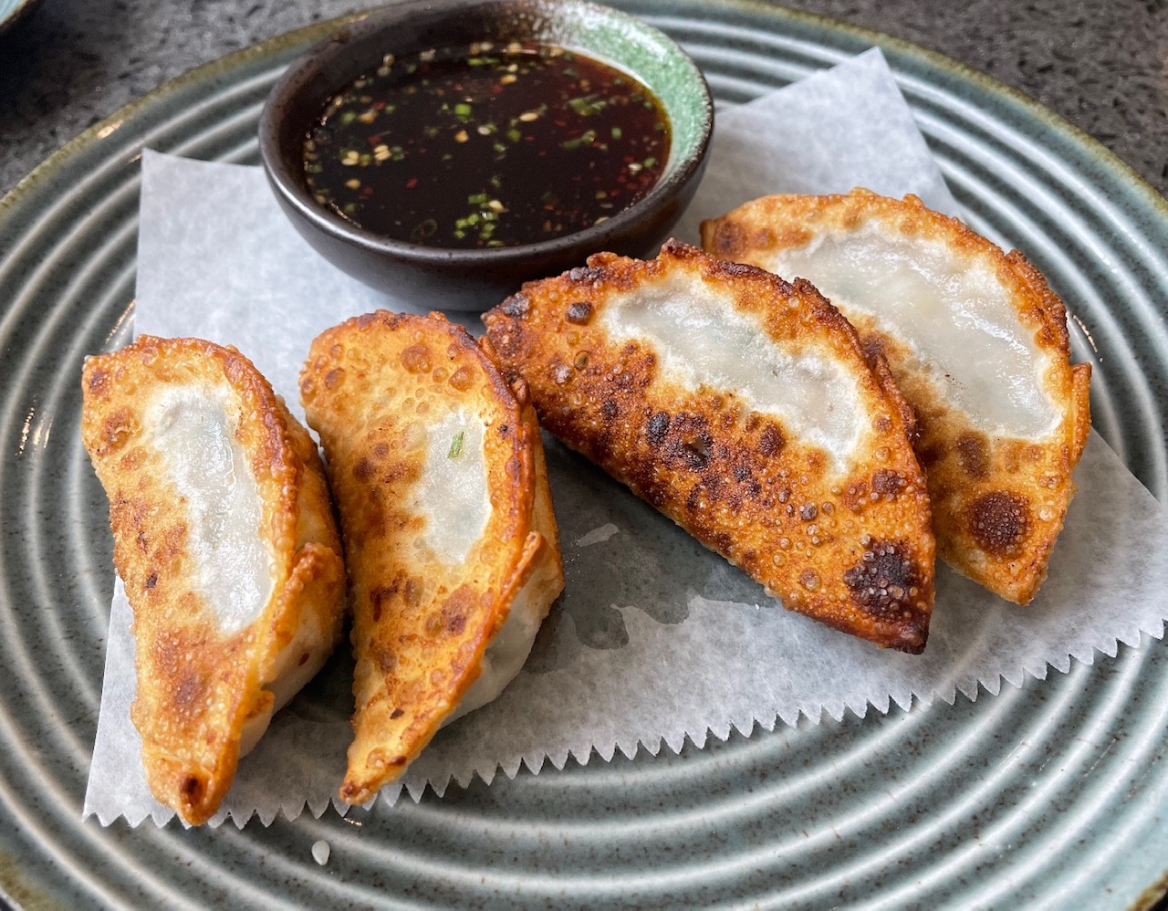 On a circular ceramic plate with white rings, there is a parchment paper with four fried dumplings and a small cup of dipping sauce. Photo by Alyssa Buckley.